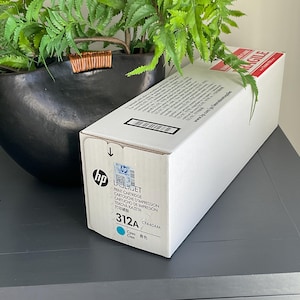 May include: A white HP LaserJet 312A ink cartridge box with a cyan color indicator. The box is labeled with product information and a barcode. The box is positioned near a black decorative pot with green plants.