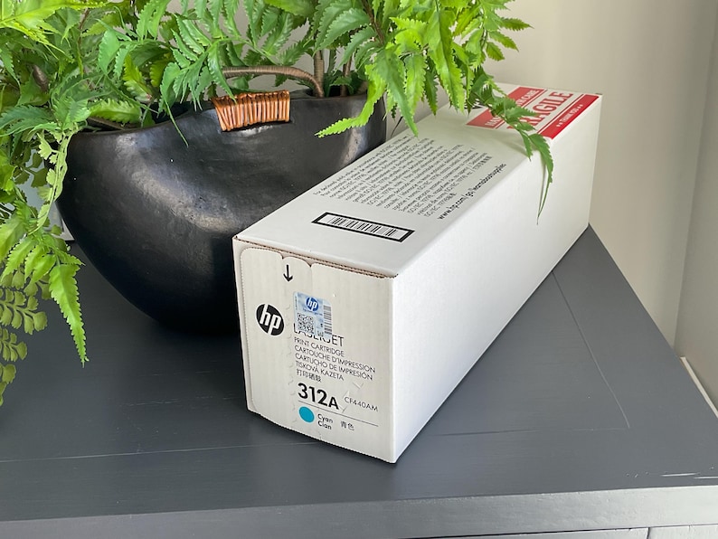 May include: A white HP LaserJet 312A ink cartridge box with a cyan color indicator. The box is labeled with product information and a barcode. The box is positioned near a black decorative pot with green plants.
