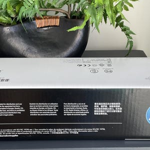 May include: A rectangular HP box with a white top and black sides. The HP logo is on the right side. The box has text in multiple languages and a recycling symbol. A warning symbol is also visible.