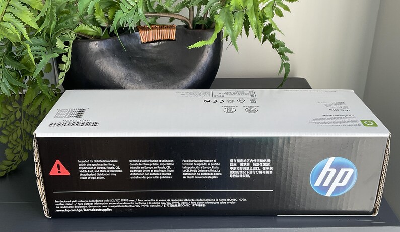 May include: A rectangular HP box with a white top and black sides. The HP logo is on the right side. The box has text in multiple languages and a recycling symbol. A warning symbol is also visible.