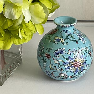 May include: A small, round, turquoise ceramic vase with a floral design in pink, blue, and yellow. The vase has a narrow neck and is displayed next to a glass vase with green and yellow flowers.