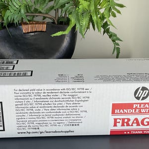 May include: A white HP box with black barcodes and text. The box has a red and white "PLEASE HANDLE WITH CARE FRAGILE" sticker. The box is on a dark surface, with a plant in the background.