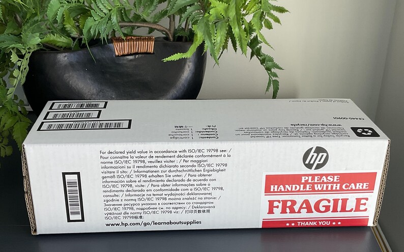 May include: A white HP box with black barcodes and text. The box has a red and white "PLEASE HANDLE WITH CARE FRAGILE" sticker. The box is on a dark surface, with a plant in the background.