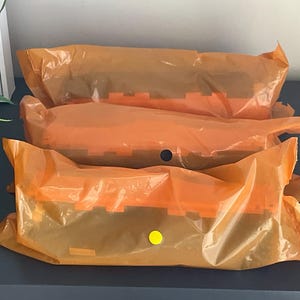 K&ouml;nnte beinhalten: Drei orangefarbene, in Kunststoff verpackte Pakete, die auf einer dunkelblauen Oberfl&auml;che gestapelt sind. Die Pakete sind mit Klebeband versiegelt und haben einen gelben Aufkleber auf einem. Die Pakete scheinen ein Produkt mit einem gekerbten Design zu enthalten.