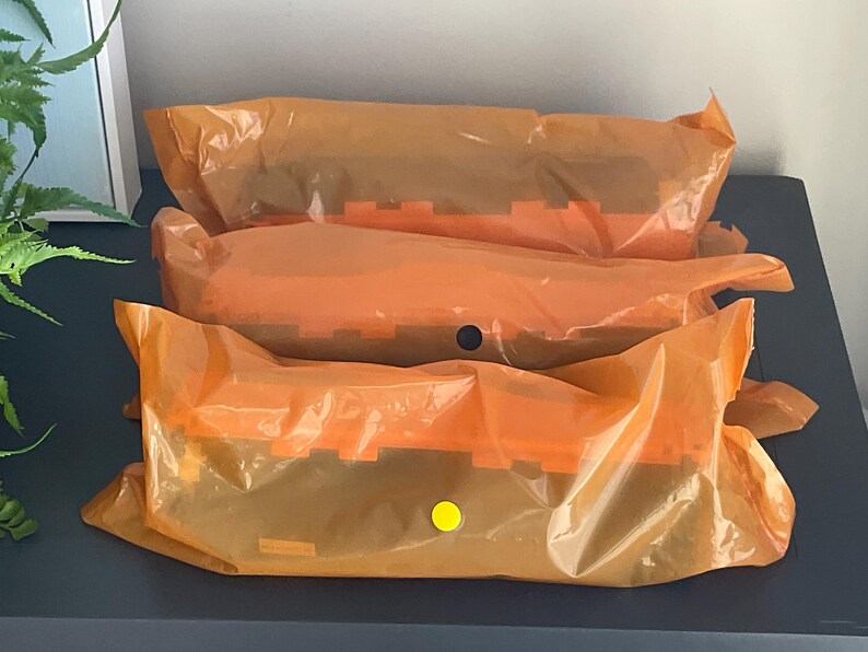 K&ouml;nnte beinhalten: Drei orangefarbene, in Kunststoff verpackte Pakete, die auf einer dunkelblauen Oberfl&auml;che gestapelt sind. Die Pakete sind mit Klebeband versiegelt und haben einen gelben Aufkleber auf einem. Die Pakete scheinen ein Produkt mit einem gekerbten Design zu enthalten.