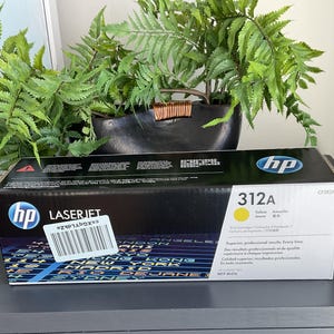 May include: Black and yellow HP LaserJet 312A toner cartridge box. The box has the HP logo and product information. The box is on a dark gray surface with a plant in the background.