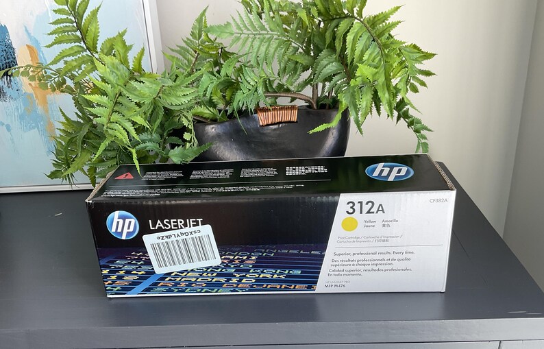 May include: Black and yellow HP LaserJet 312A toner cartridge box. The box has the HP logo and product information. The box is on a dark gray surface with a plant in the background.