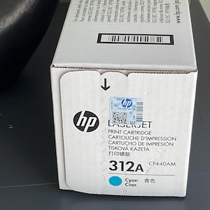 May include: A white HP LaserJet print cartridge box with the HP logo and the text "312A Cyan". The box has a blue and white sticker and a black arrow indicating the opening direction.