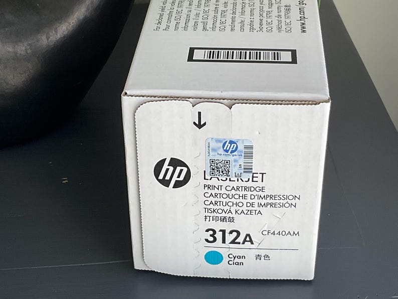 May include: A white HP LaserJet print cartridge box with the HP logo and the text "312A Cyan". The box has a blue and white sticker and a black arrow indicating the opening direction.