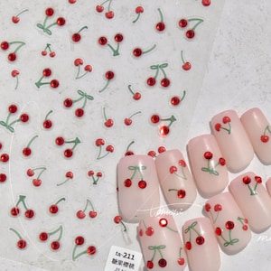 May include: A set of false nails with a cherry design. Each nail is painted a light pink color and has a small cherry with a green stem and red fruit. The cherries have a single rhinestone in the center. The nails are arranged in a row on a white background. The image also includes a sheet of nail stickers with a cherry design.