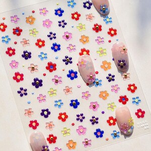 May include: A sheet of colourful flower nail stickers with a clear background. The stickers are small and feature a variety of colours, including red, blue, yellow, green, and purple. The stickers are arranged in a grid pattern and are perfect for adding a touch of whimsy to your nails.