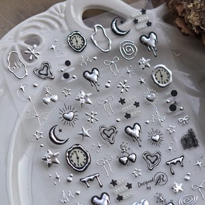 May include: A sheet of silver and black sticker decals featuring various designs, including hearts, stars, moons, clocks, and swirls. The sheet is labeled "Designed by R".