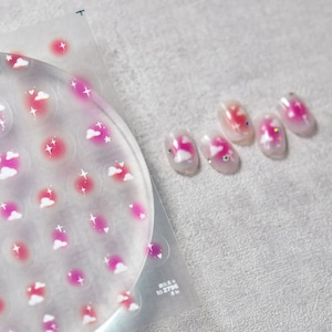 May include: A sheet of nail stickers with a pink and white cloud and star design. The sheet is clear and has a circle of stickers in the center. There are five nails with the same design on a white surface.