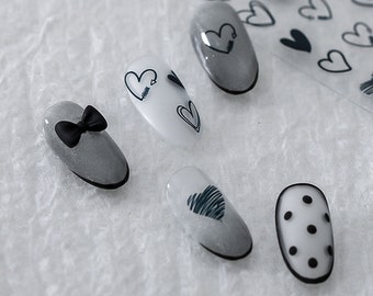 Pode incluir: Cinco designs de unhas diferentes com corações pretos e brancos, um laço e bolinhas.