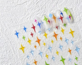 May include: Close-up of five clear nail tips with colourful star designs. The nail tips are arranged in a row on a white background. The background is covered in small, colourful star stickers.