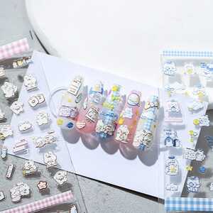 May include: Clear nail art stickers featuring cartoon bears, stars, clouds, and other cute designs. The stickers are applied to long, clear nails with various pastel-colored bases and patterns. A sheet of stickers with a pink and white checkered border is also visible.