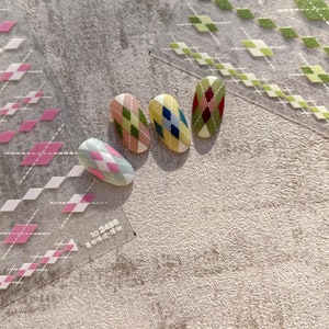 Puede incluir: Cuatro pegatinas ovaladas para uñas con un patrón de cuadros. Las pegatinas son en tonos de verde, amarillo, rosa y rojo. Las pegatinas están dispuestas en una fila sobre una superficie gris texturizada. El texto "TO 2486" y "美甲贴纸" es visible en una pegatina blanca en la esquina inferior izquierda de la imagen.