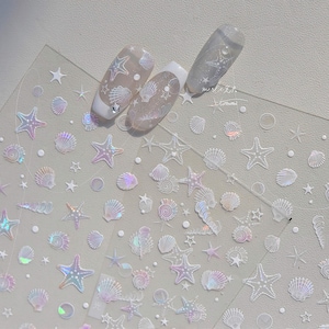 Pode incluir: Autocolantes transparentes para nail art com estrelas do mar, conchas e estrelas iridescentes. Os autocolantes têm um efeito holográfico, e alguns são aplicados em unhas cuidadas. O tema geral é praia ou oceano.