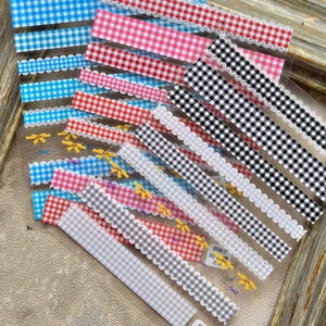 May include: A sheet of transparent adhesive tape with various gingham patterns in red, blue, black, and white. The tape also features a white lace trim and yellow bows.