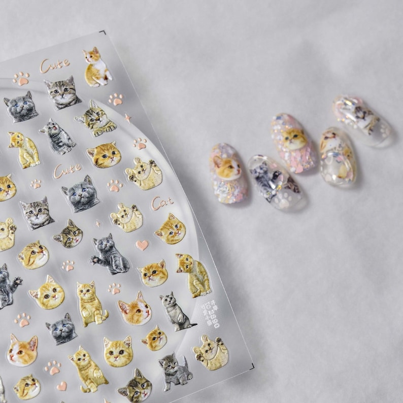 May include: A sheet of clear nail stickers with images of cute cats. The stickers are arranged in rows and columns, and the cats are in various poses. The sheet is labeled "Cute Cat" and has a pink paw print design. The image also shows five fingernails with the cat stickers applied.