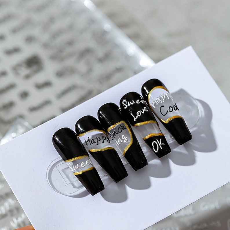 May include: Set of six black press-on nails with gold accents and white lettering. The nails feature the words "Sweet Love", "Happy", "Cod", "Happy Smoking", "ing", and "OK".