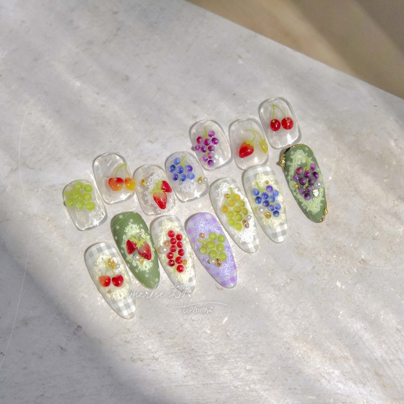 Pu&ograve; includere: Un set di 12 unghie finte con vari design di frutta. Le unghie presentano ciliegie, fragole e uva con foglie verdi e dettagli dorati. Le unghie sono disposte su uno sfondo a scacchi bianco e grigio.