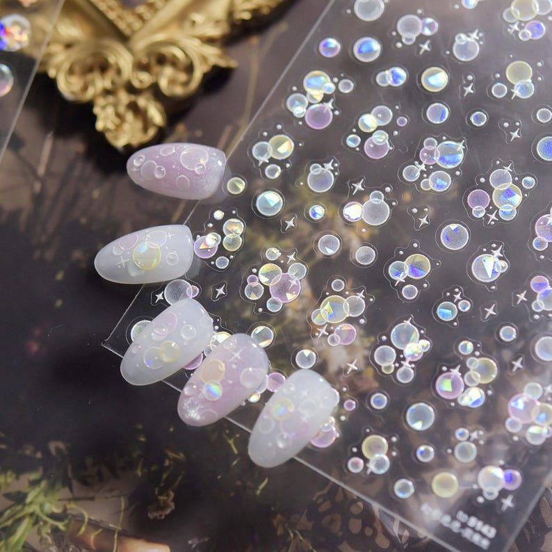 May include: A sheet of iridescent nail art stickers featuring bubble and star designs. Several manicured nails are shown with the stickers applied, showcasing a pearlescent effect. The stickers are clear with a holographic finish.