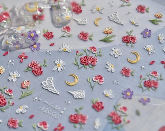 Puede incluir: Hoja de pegatinas de arte de uñas con diseños florales, incluyendo rosas rojas, margaritas blancas, flores moradas y lunas crecientes doradas. El fondo es de color azul claro.