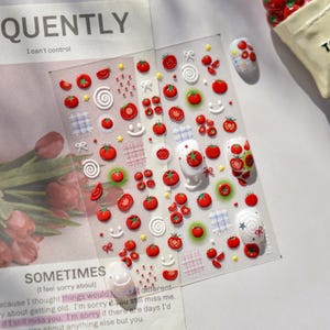 May include: Clear sheet of nail art stickers featuring red tomato designs, white swirls, and smiley faces. The sheet includes various sizes of tomatoes and decorative elements. The text "QUENTLY" and "SOMETIMES" are visible.