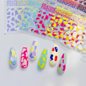 Op de afbeelding: Zes kunstnagels met een koe-print ontwerp. De nagels zijn in verschillende kleuren geverfd, waaronder roze, blauw, geel en groen. Elke nagel heeft een kleine zilveren stud.