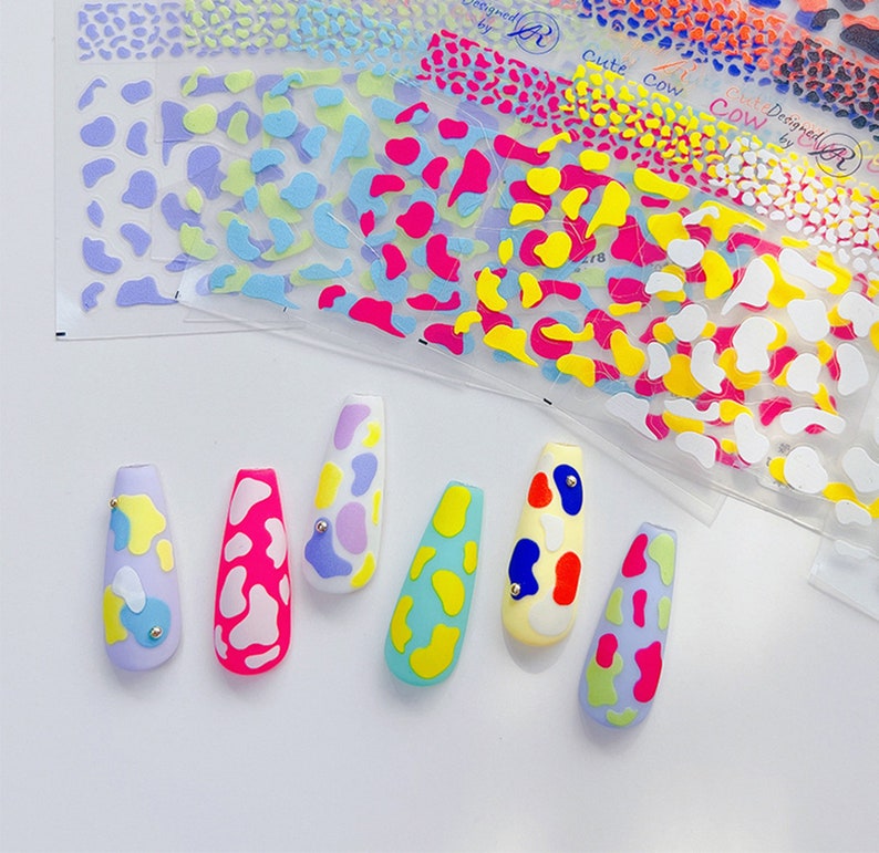 Op de afbeelding: Zes kunstnagels met een koe-print ontwerp. De nagels zijn in verschillende kleuren geverfd, waaronder roze, blauw, geel en groen. Elke nagel heeft een kleine zilveren stud.