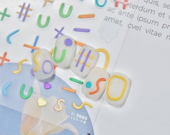 May include: A set of white nail stickers with colourful designs. The designs include letters, lines, and geometric shapes. The stickers are arranged in a row on a white surface.