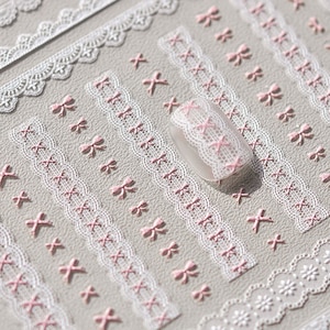May include: White lace nail stickers with pink bows. The stickers are arranged in rows on a gray background.