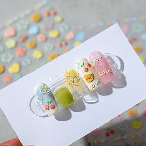 May include: Five fake nails with different designs. The nails are decorated with butterflies, cherries, a bear, and the words "good" and "love".