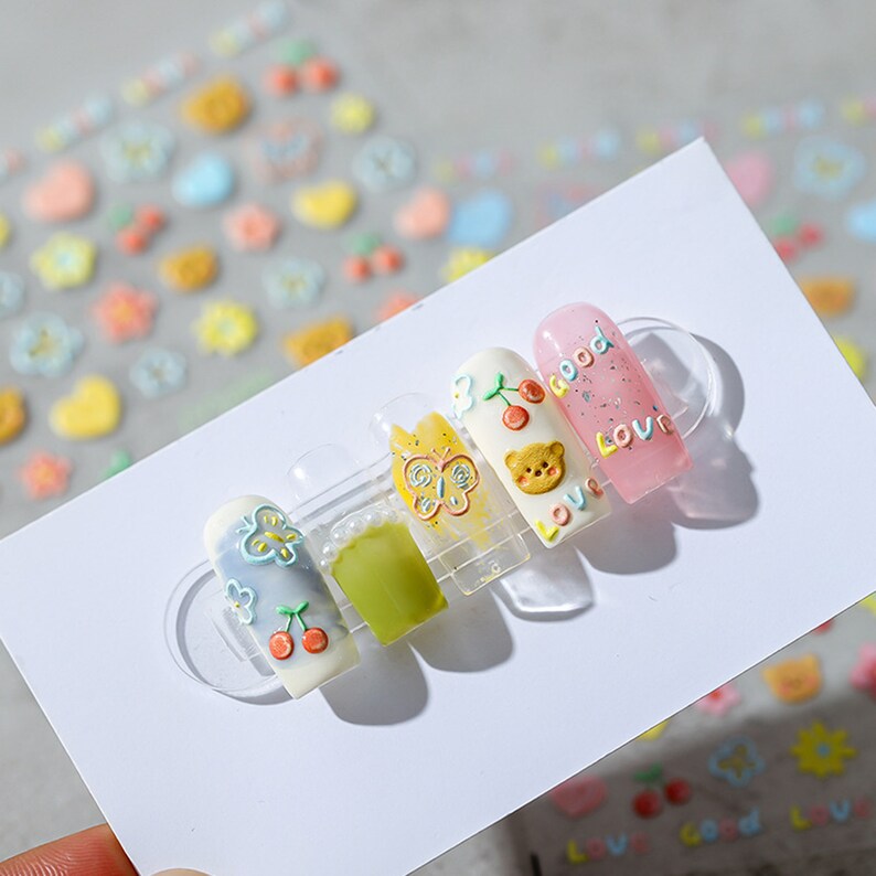 May include: Five fake nails with different designs. The nails are decorated with butterflies, cherries, a bear, and the words "good" and "love".