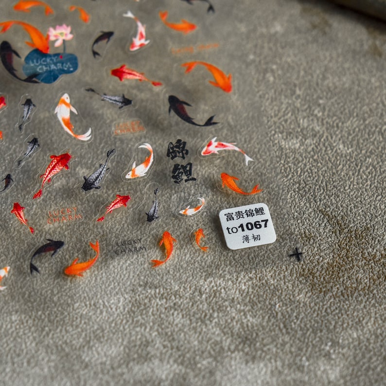 May include: A sheet of clear stickers with koi carp designs in orange, red, black, and white. Some stickers have the text "Lucky Charm" and Chinese characters. A small white square with text is also visible.