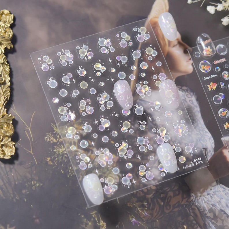 May include: Clear nail art stickers with iridescent bubble designs. The stickers are applied to several manicured nails. The image also shows a second sheet of stickers with a goldfish design and the text "GOUPER" and "Carpe Diem."