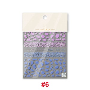 Op de afbeelding: Een pakje nagelkunst stickers met een koe-print ontwerp in tinten paars en blauw. De stickers zijn gelabeld "cute cow" en worden geleverd in een doorzichtige plastic zak met een wit label waarop "nailart" staat.