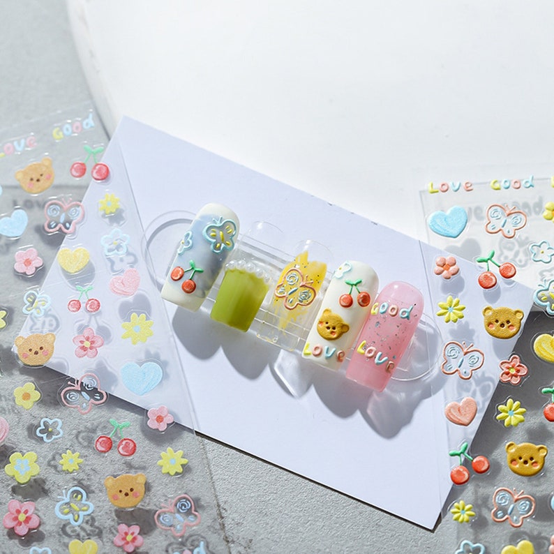 May include: A sheet of nail art stickers with a variety of cute designs, including butterflies, hearts, cherries, flowers, and bears. The stickers are on a clear background and are ready to be applied to nails.