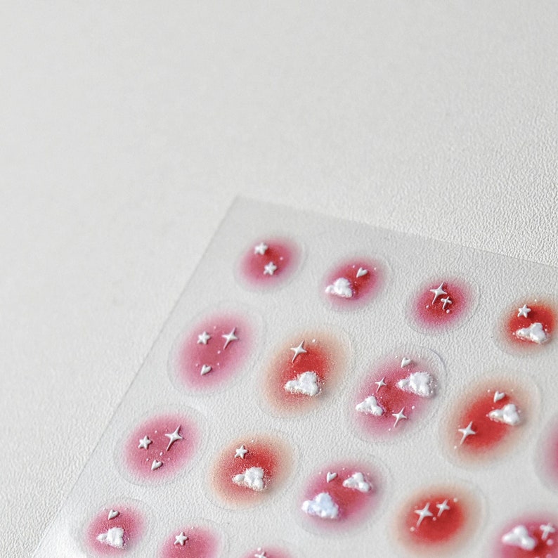 May include: A sheet of pink and white cloud and star stickers. The stickers are arranged in a grid pattern on a clear sheet.