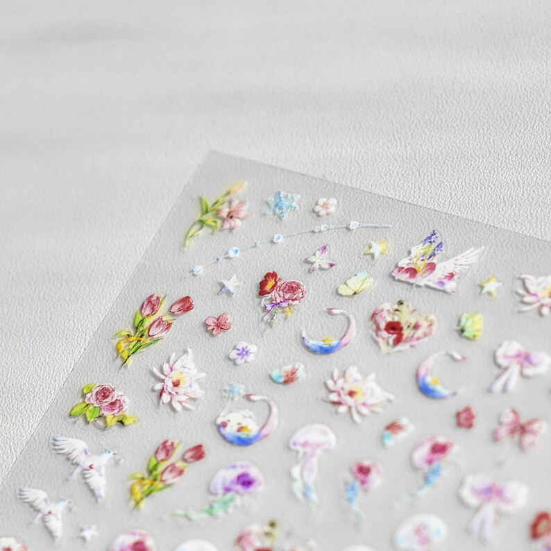 May include: A sheet of clear nail stickers with various designs, including flowers, stars, moons, and butterflies. The stickers are in shades of pink, blue, green, and white.