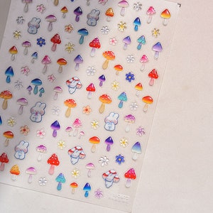 May include: A sheet of colorful stickers featuring mushrooms, flowers, and bunnies. The stickers are glossy and have a 3D effect. The sheet is white with a light pink background.