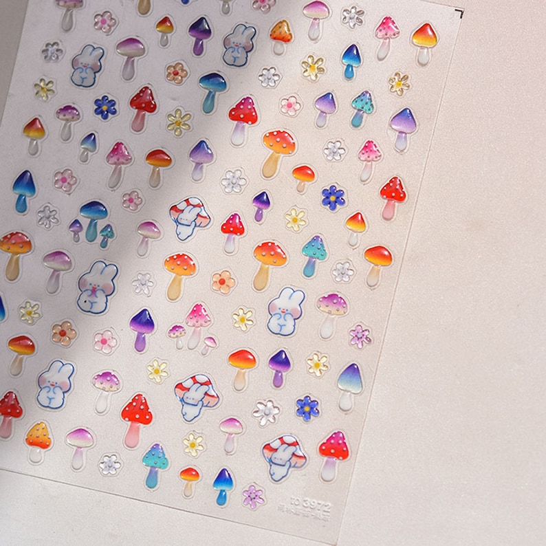 May include: A sheet of colorful stickers featuring mushrooms, flowers, and bunnies. The stickers are glossy and have a 3D effect. The sheet is white with a light pink background.