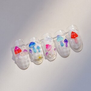 May include: Five clear fake nails with a checkered pattern and mushroom designs. The mushrooms are in various colors, including red, blue, pink, and purple.
