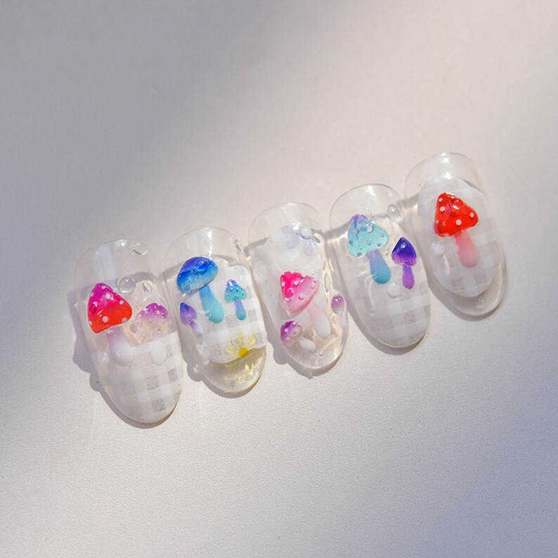 May include: Five clear fake nails with a checkered pattern and mushroom designs. The mushrooms are in various colors, including red, blue, pink, and purple.