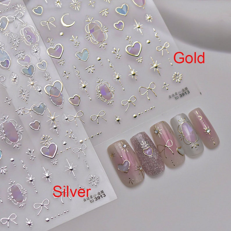 May include: Two sheets of nail art stickers with a variety of designs, including hearts, stars, bows, and moons. The stickers are available in gold and silver. The image also shows five fingernails with the stickers applied.