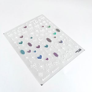 May include: A sheet of white nail stickers with a variety of designs, including hearts, stars, bows, and moons. The stickers have a holographic iridescent finish.