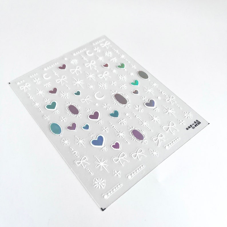 May include: A sheet of white nail stickers with a variety of designs, including hearts, stars, bows, and moons. The stickers have a holographic iridescent finish.