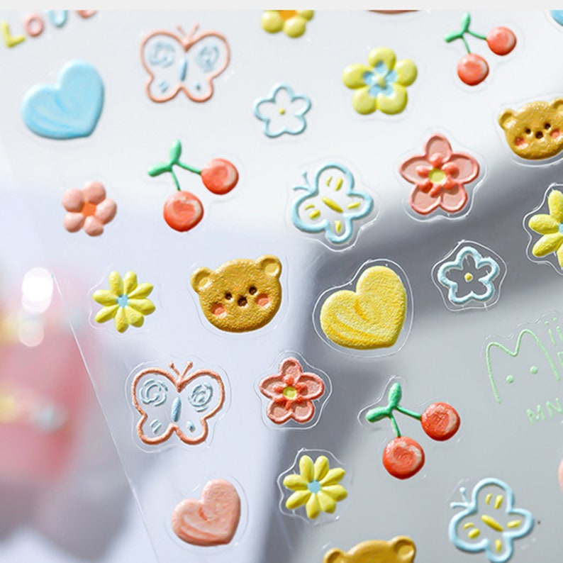 May include: A sheet of colorful, glossy stickers featuring various cute designs, including butterflies, hearts, flowers, cherries, and bears. The stickers are perfect for adding a touch of whimsy to journals, planners, and other craft projects.