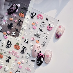 May include: Halloween-themed nail art stickers featuring pumpkins, cats, ghosts, and spiderwebs. The stickers are in black, orange, pink, and white. The image also shows several painted nails with Halloween designs.